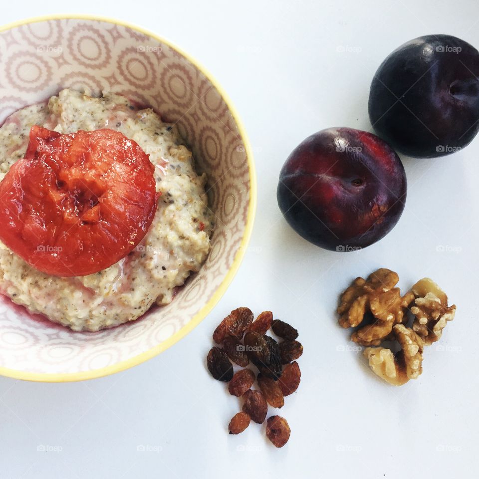 Plums raisins and walnuts around a patterned bowl containing oatmeal porridge with a stewed plum on top 