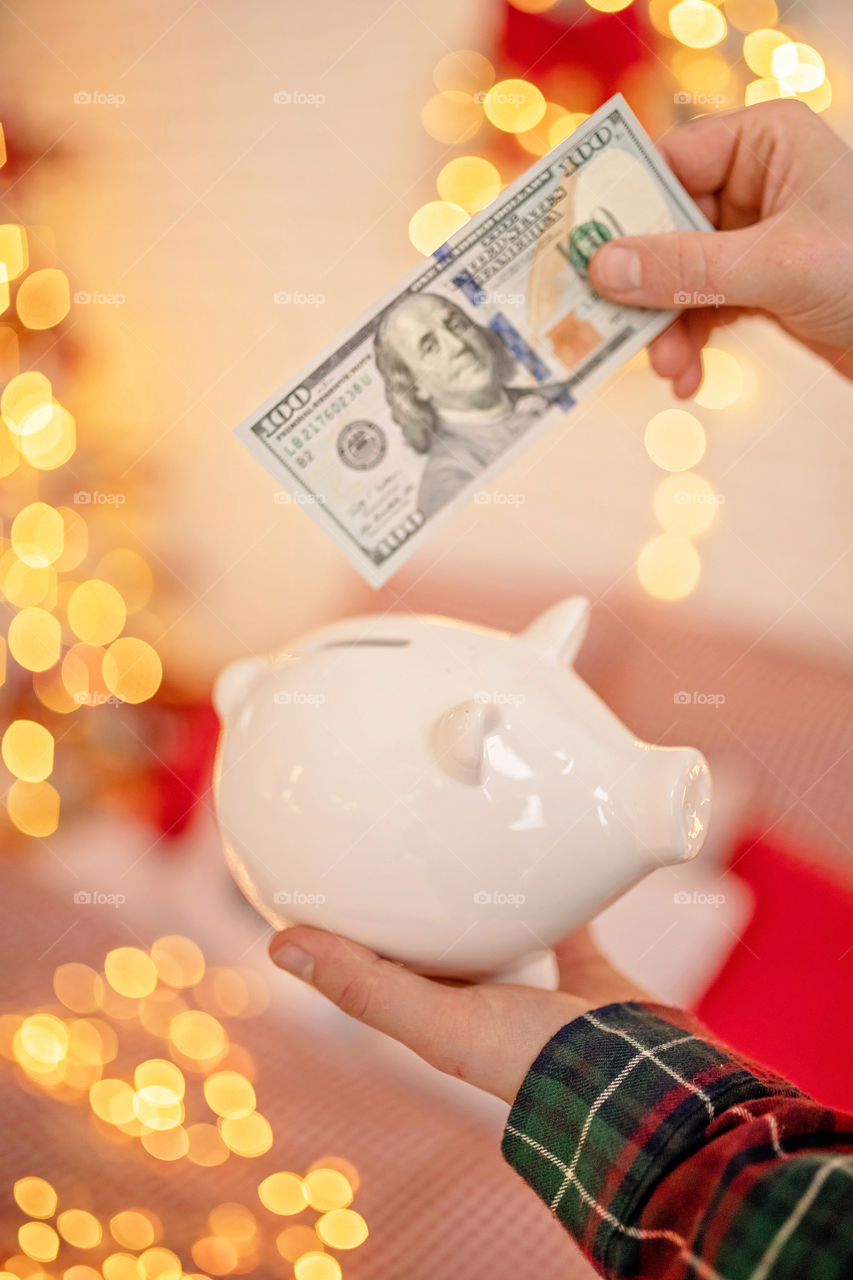 piggy bank with 100 dollars banknote in festive New Year atmosphere of scenery.  Magical bokeh with Christmas tree and bright lights.