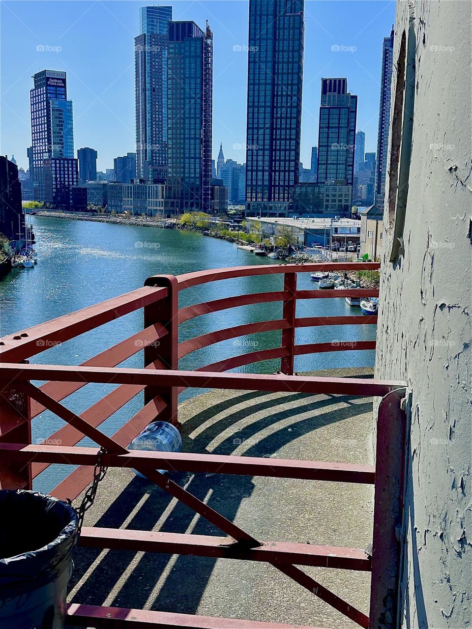 This is the center outpost of the “Pulaski Bridge” at “Newtown Creek” that connects “Greenpoint”, Bklyn to LIC. Across the “East River” we see “Manhattan”. The afternoon sun casts dramatic shadows onto the asphalt platform. 2024. Hypnotic Productions