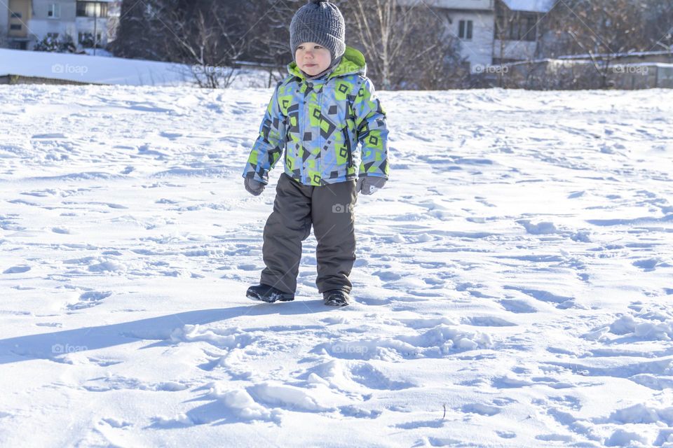A child with a serious expression on his face in winter clothes jackets, pants, hat and boots in winter on the white snow on the street and in the park in nature plays winter fun.