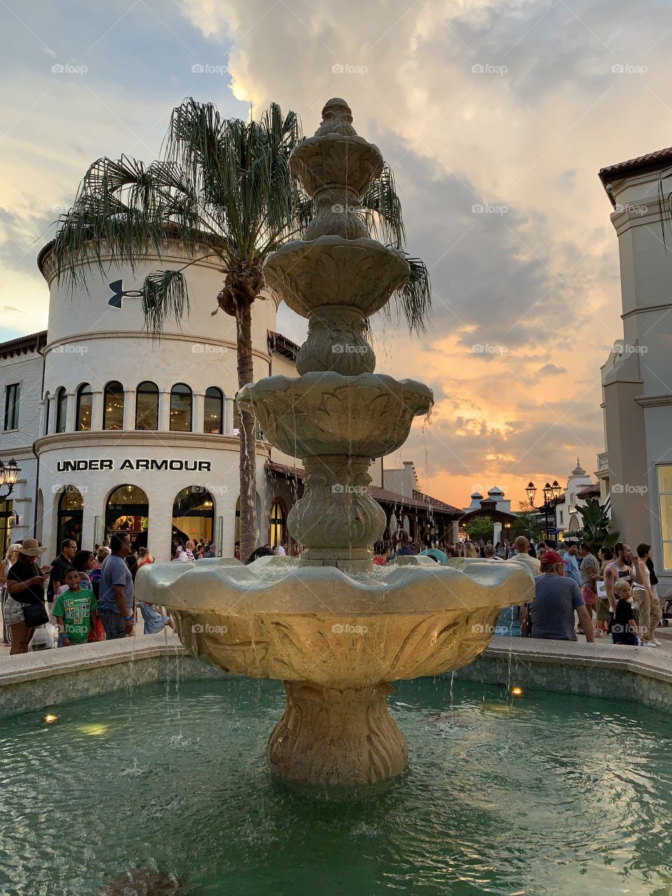European style plaza with shops, fountain, palm tree, people and sunset
