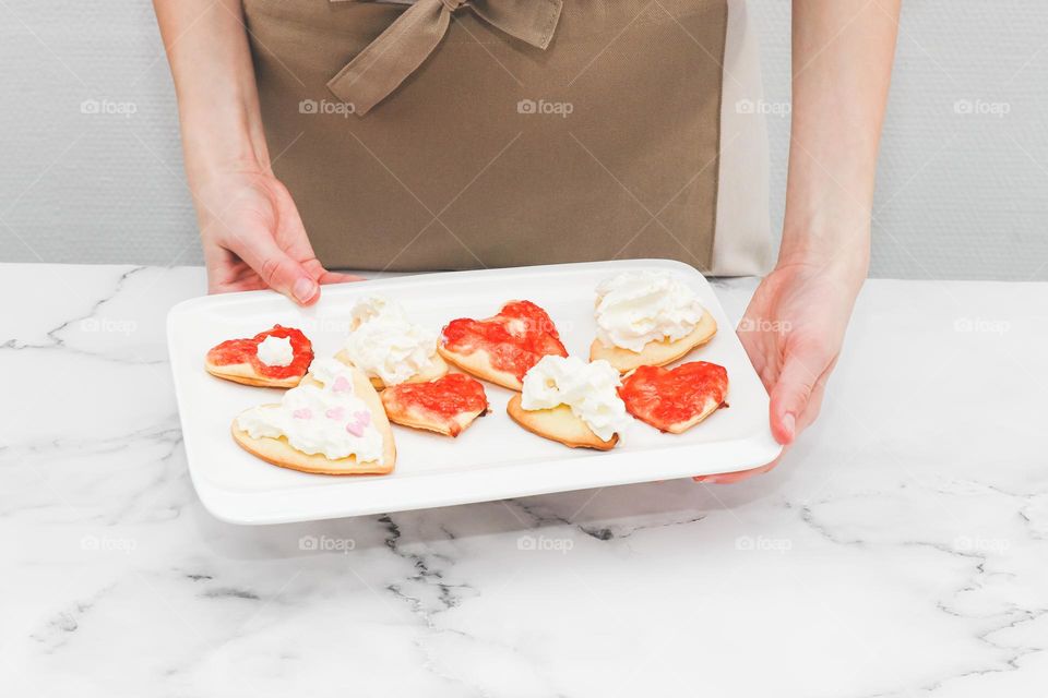 Caucasian teenage girl in an apron holds in her hands a rectangular dish with homemade heart-shaped cookies with raspberry jam. whipped cream standing at a marble table against a white wall, close-up side view.