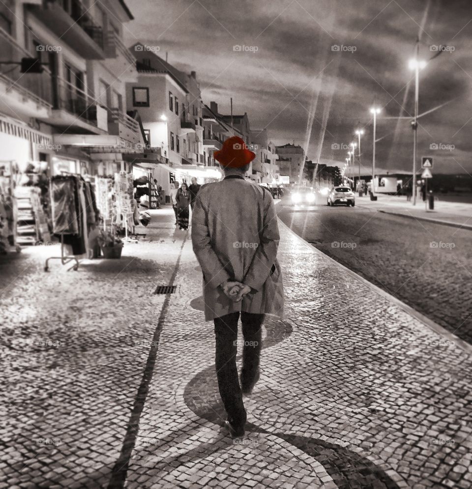 Man walks along the cobbled street in Nazaré. It is nighttime, the shops are still open and the street lights are on. The image is black and white apart from the man’s red hat