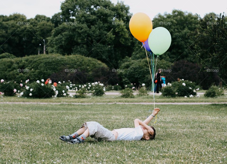 A little blond boy in a white T-shirt lies on a green field, holding a bunch of colorful balloons in his hand, back view 