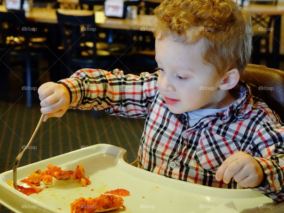 A little boy sitting in a high chair digs into his favorite snack of pepperoni pizza with great vigor. 