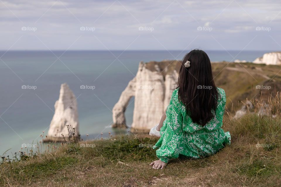 One young caucasian brunette girl with flowing hair in a green dress sits from the back on a mountain top enjoying the view of the north sea and a natural arch in Étretat, Normandy, France, close-up side view.