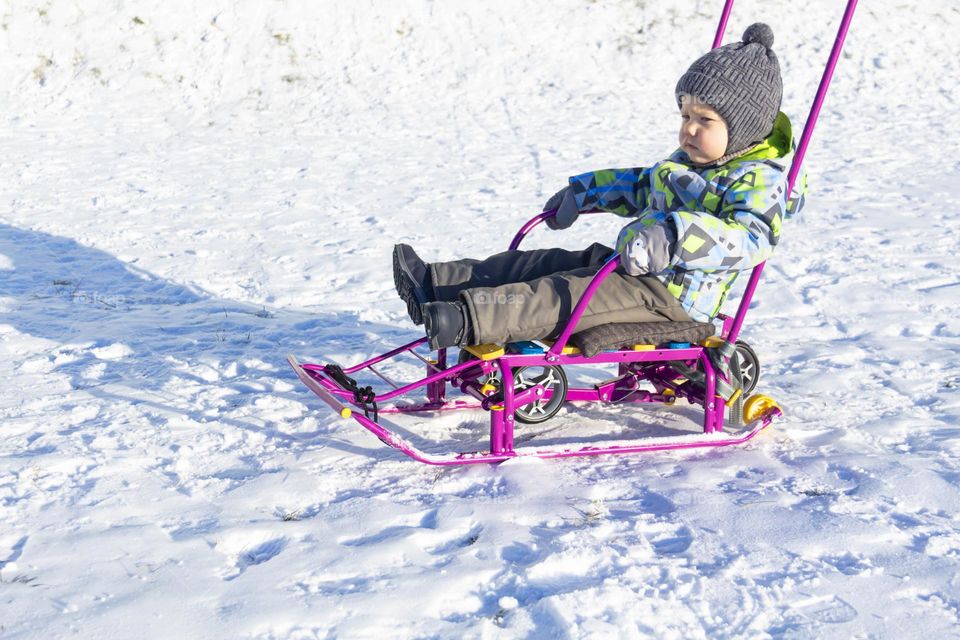 A child with a serious expression on his face in winter clothes jackets, pants, hat and boots in winter on white snow on the street and in the park in nature sledding and playing winter fun.