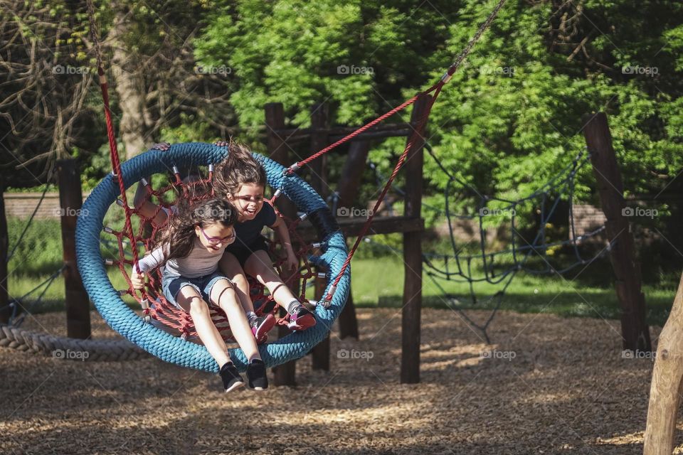 Two beautiful happy caucasian brunette girls with laughing out loud ride together on a round hanging swing on a clear sunny day in a public park on a playground, close-up side view.