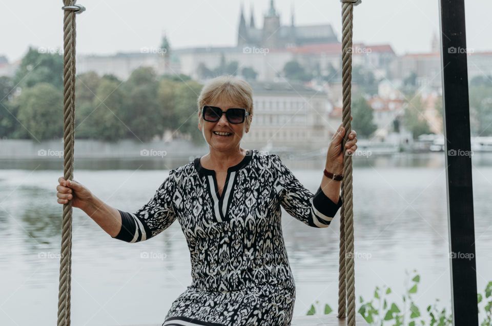 Portrait of one beautiful Caucasian elderly blonde woman in dress and sunglasses with happy smile riding on swing on summer day against background of blurred river and city buildings, side view close up.