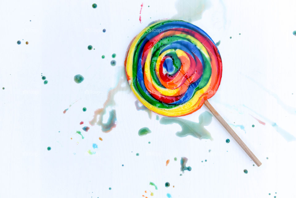 Flat lay of a rainbow swirl lollipop surrounded by colored sugar splatter on a white backdrop