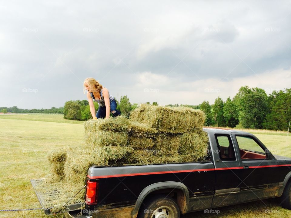 A young farm girl stacks hay bales into her old trusty truck for her horses before a coming storm