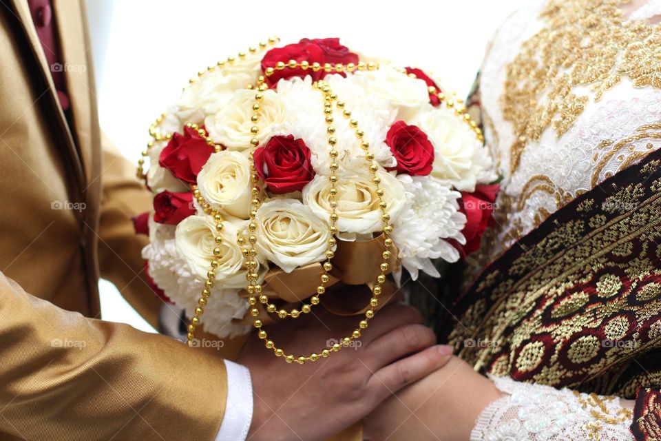 Man and woman holding a bouquet of rose red and white flowers while holding hands in wedding.