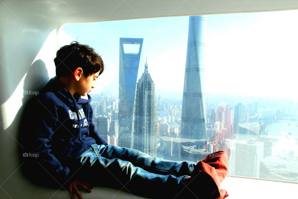 A cute 4 years old Little boy contemplates the view of PuDong skyscrapes, sitting at the windows of the Space Module from the Pearl Tower in Shanghai, 370 meter High, 2015