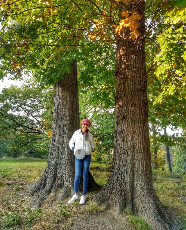 A woman wearing a red hat, white sweater. Hands on her pocket, standing with two big trees background. 