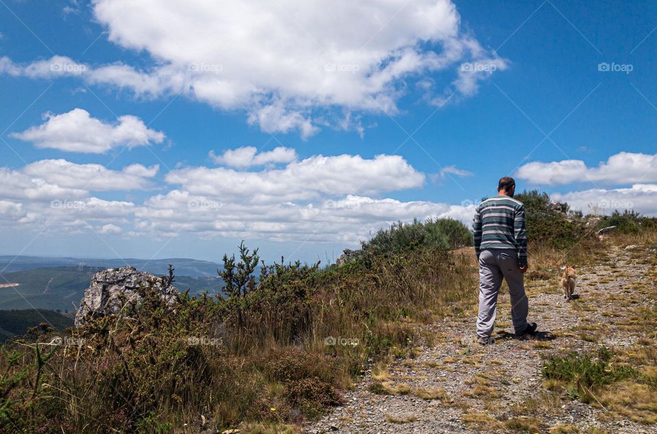 A man and his dog walk up a trail alongside a large boulder under a blue sky with fluffy white clouds