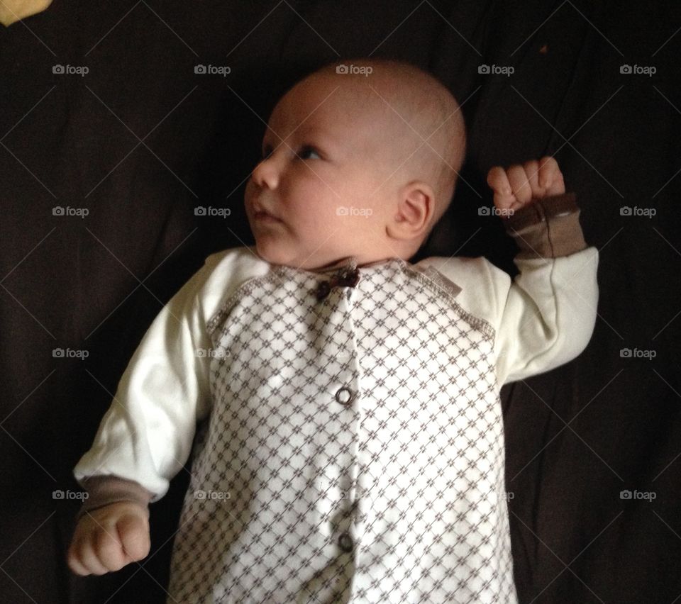 A newborn baby boy lying on a dark bed, with one hand up with a fist and the other down. Dressed in a sleepsuit with a bow.