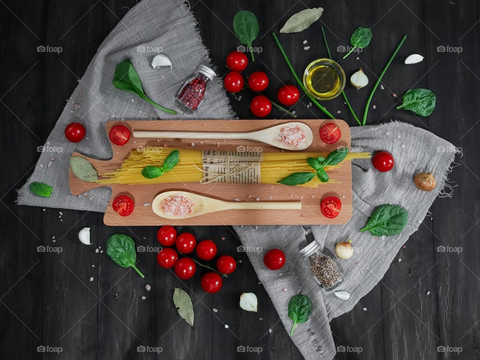 Raw spaghetti tied with jute thread on a cutting board with wooden spoons, tomatoes, basil leaves, salt, seasonings and olive oil on a black wooden yone, flat lay close-up.