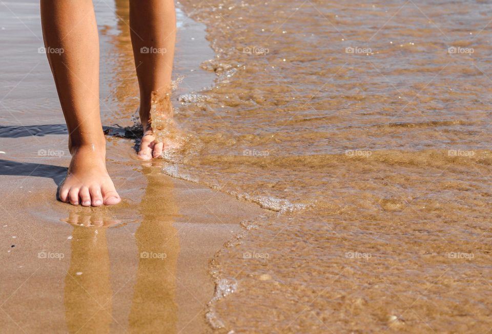 A beautiful view of children's feet walking along the sandy shore with an incoming sea wave on them from the right side copy space, close-up side view.