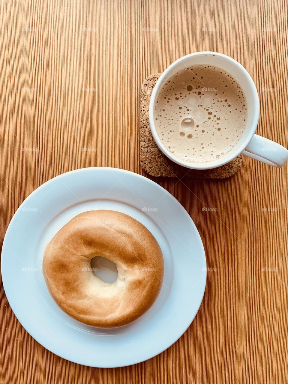 Hot beverage mug and bagel on white ceramic dish seen from above on wooden surface 