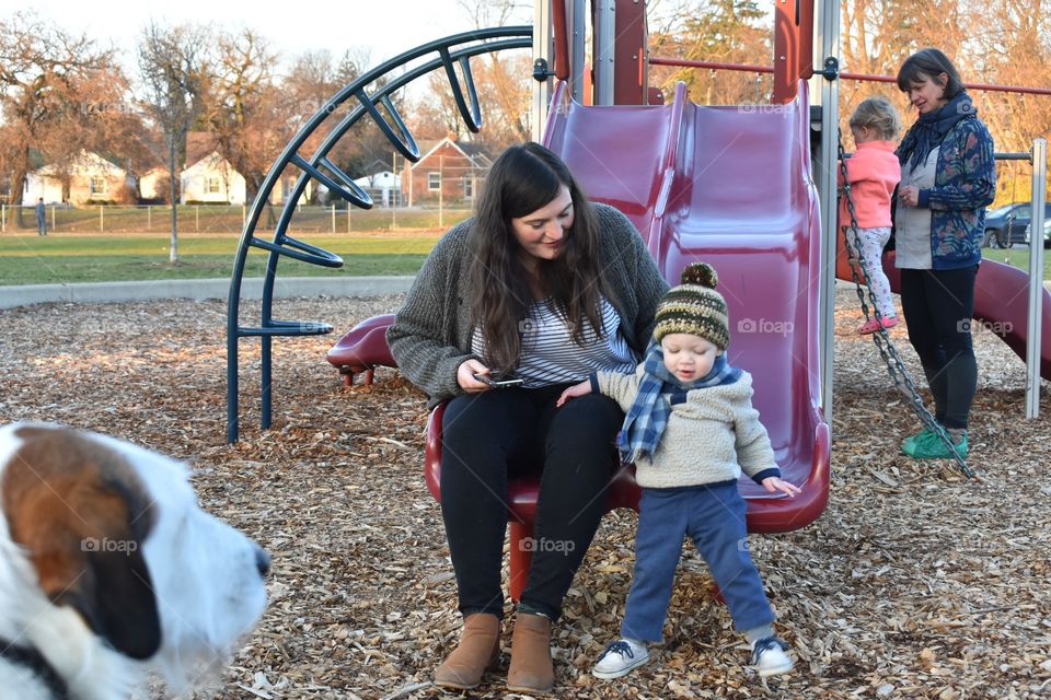 Cute toddler boy playing at playground with his mother as his dog watches 