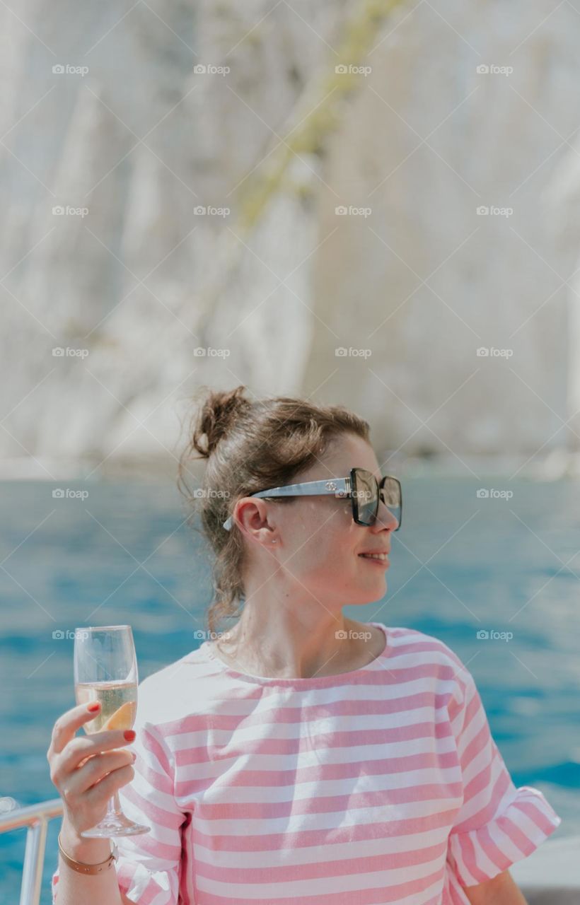 Portrait of one young beautiful Caucasian brunette girl in sunglasses, in a pink dress with a glass of champagne in her hand, sitting on the stern of a boat on a sunny summer day on blurred rocks and the sea, close-up side view.