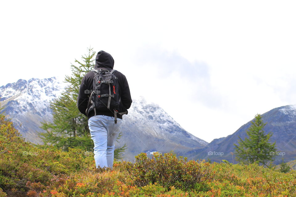 A man in blue jeans, a sweatshirt with a hood and a backpack walks along the Alpine mountains