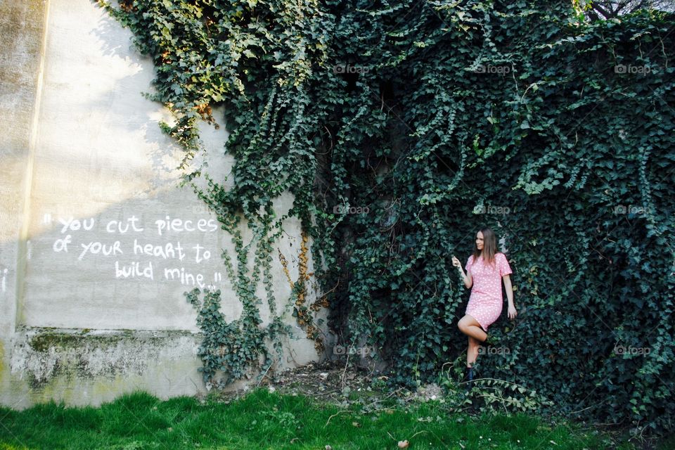 Woman in pink dress standing against poison ivy wall looking at love qoute written on the wall