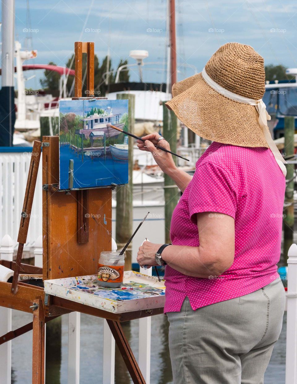 woman with large sun hat standing at marina with easel and painting the scene that she is looking at