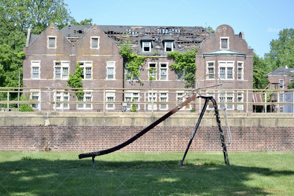 A sideways view of an old metal slide in front of an old abandoned brick building that’s overgrown with vegetation 