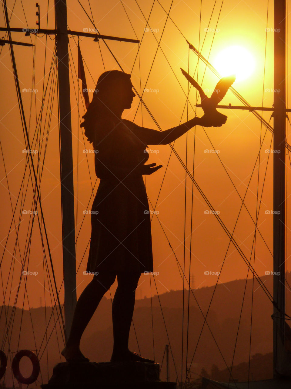 A statue of a lady holding a bird up towards the sunset 