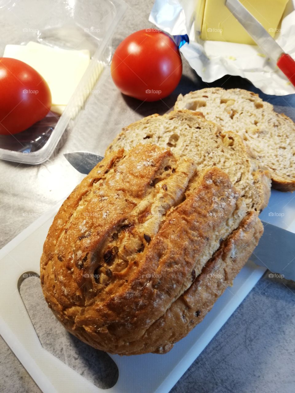 A loaf of bread with fruit and seeds on a chopping board, some crumbs on it. An edge of a knife is wavy. In an open butter package is another knife with red handle. A tomato and cheese slices in a plastic box