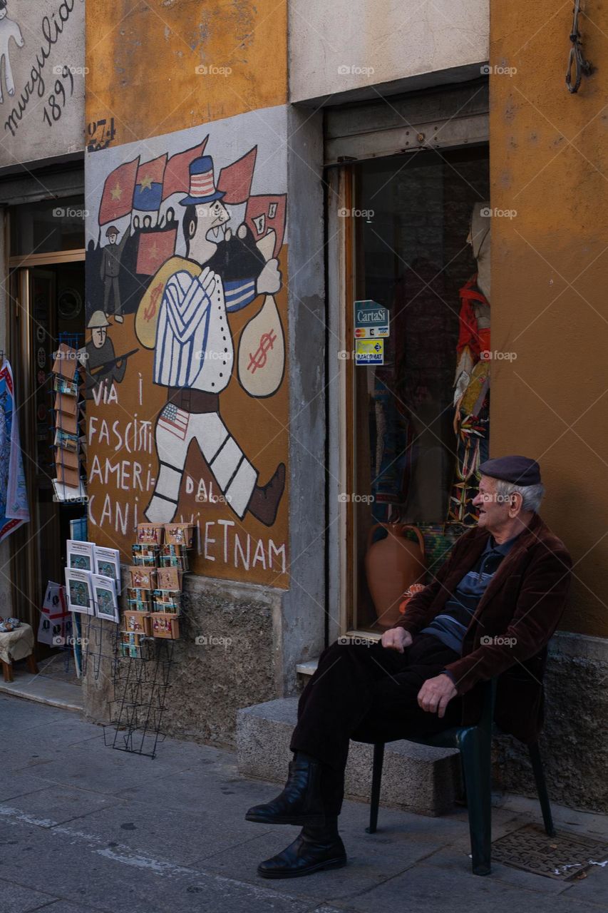 Old man sitting beside a souvenirs shop and a mural painting in Orgosolor - Sardinia
