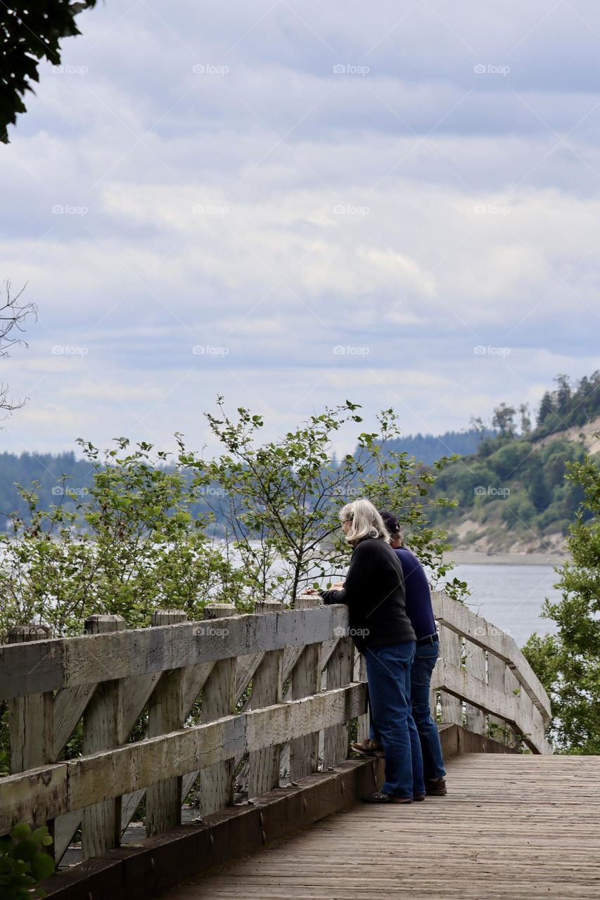 A mature man and woman stand together on a wooden bridge, it’s path leading to the shoreline of the Puget Sound in Washington State 