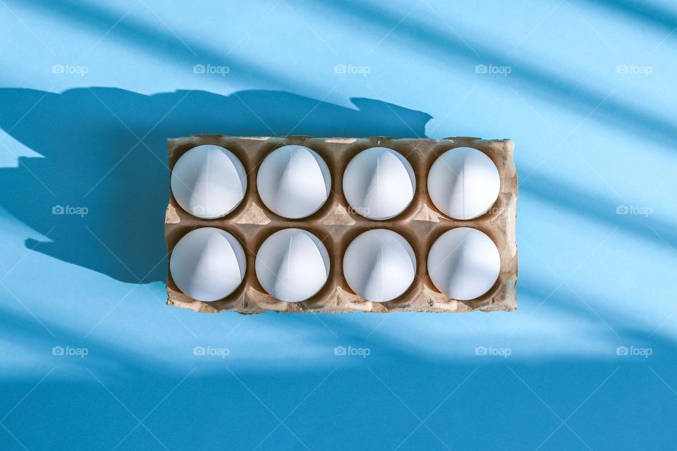 One tray with eight eggs lies in the center on a blue background with geometric shadows, flat lay close-up.