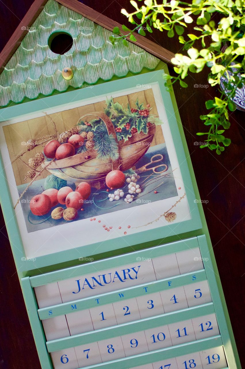 Green wooden perpetual calendar with interchangeable decorative image, ivory-colored date tiles with light blue lettering, and a small plant partially illuminated by sunlight on a dark wood surface 