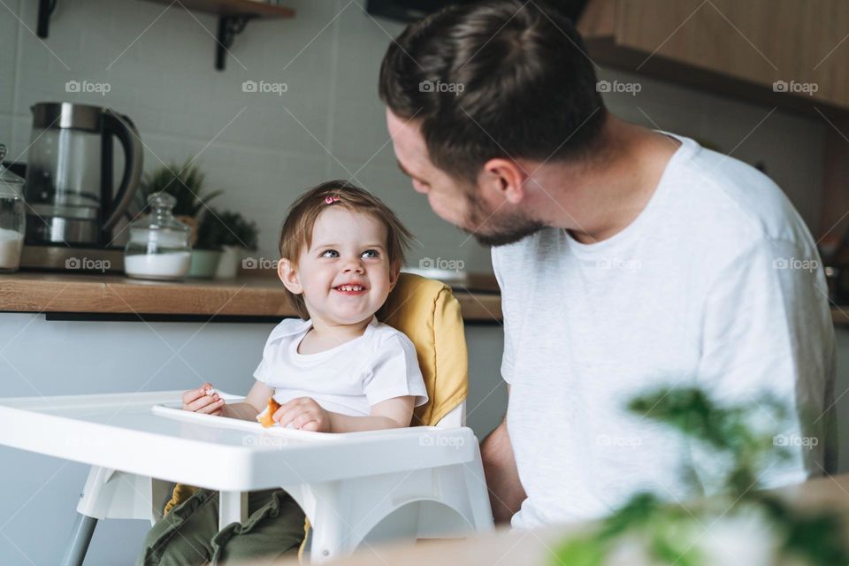 Happy father young man feeds baby girl little daughter in kitchen at home