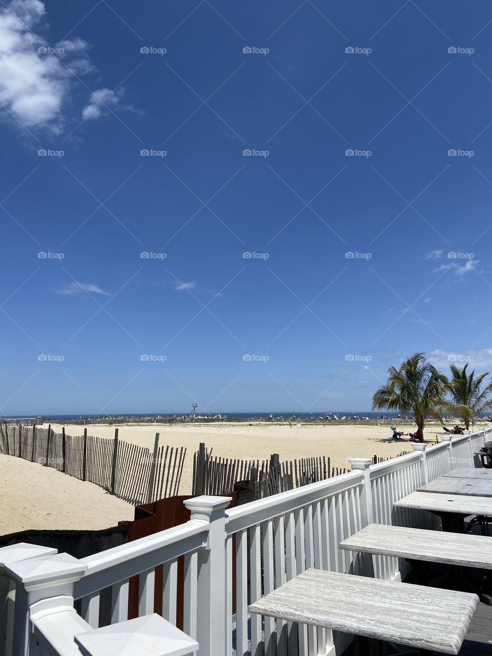 Bench fence forms a triangle of sandy beach with the ocean on one edge beneath bright blue sky. A little patch of summer paradise with people lounging beneath the palm trees in the distance.