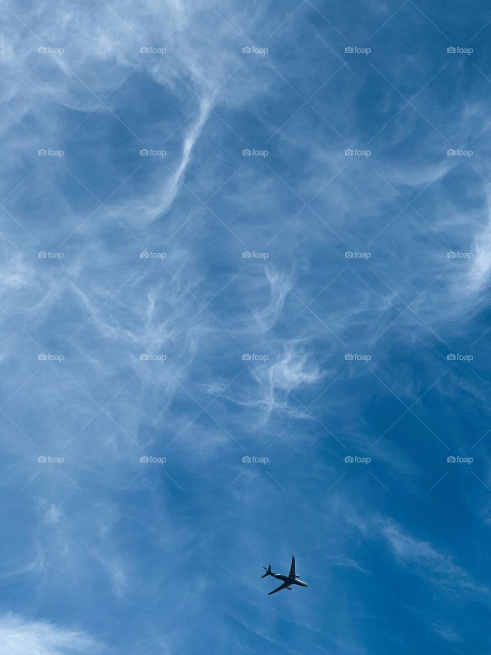 An airplane soars across a blue sky, trailed by soft, wispy clouds.