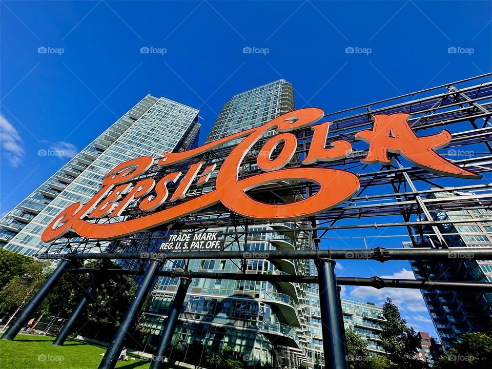 The “PEPSI COLA” sign is located in a city park on the shore of the “East River” near “Newtown Creek” in LIC, Queens. Its shapely elegant letters wind their way around the billboard contrasting in red against the blue sky. 2024. Hypnotic Productions
