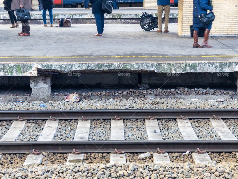 Feet and legs of travelers or commuters waiting for their train at the platform of a railway station showing no faces but part of the railroad tracks