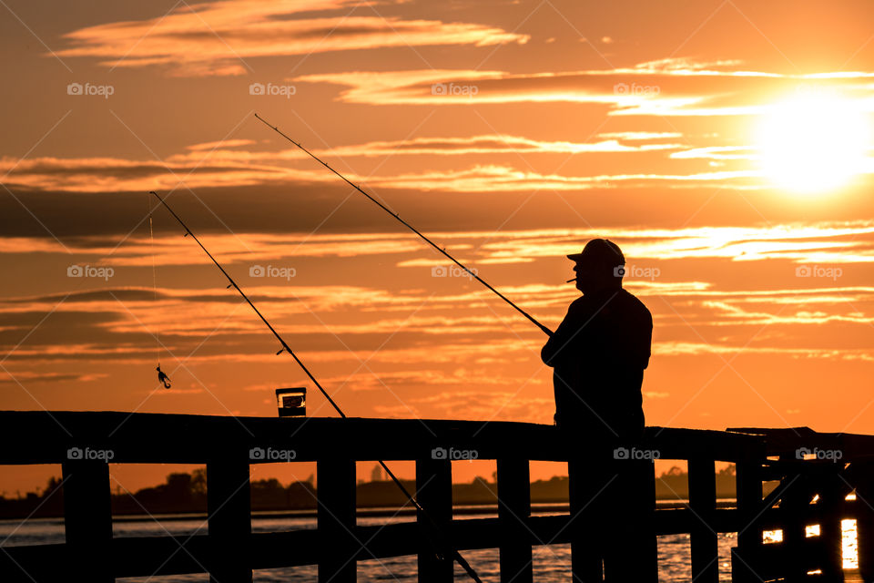 Silhouette of a man standing on a fishing pier during a vibrant sunset with two fishing poles and a jar of bait. 