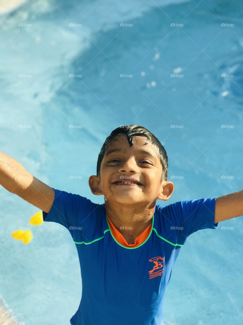 A 6 year old boy enjoying a lot at swimming pool with cheerful face in summer and he is wearing blue colour swimsuit and he looks upside towards the sky by spreading hands both sides ๐