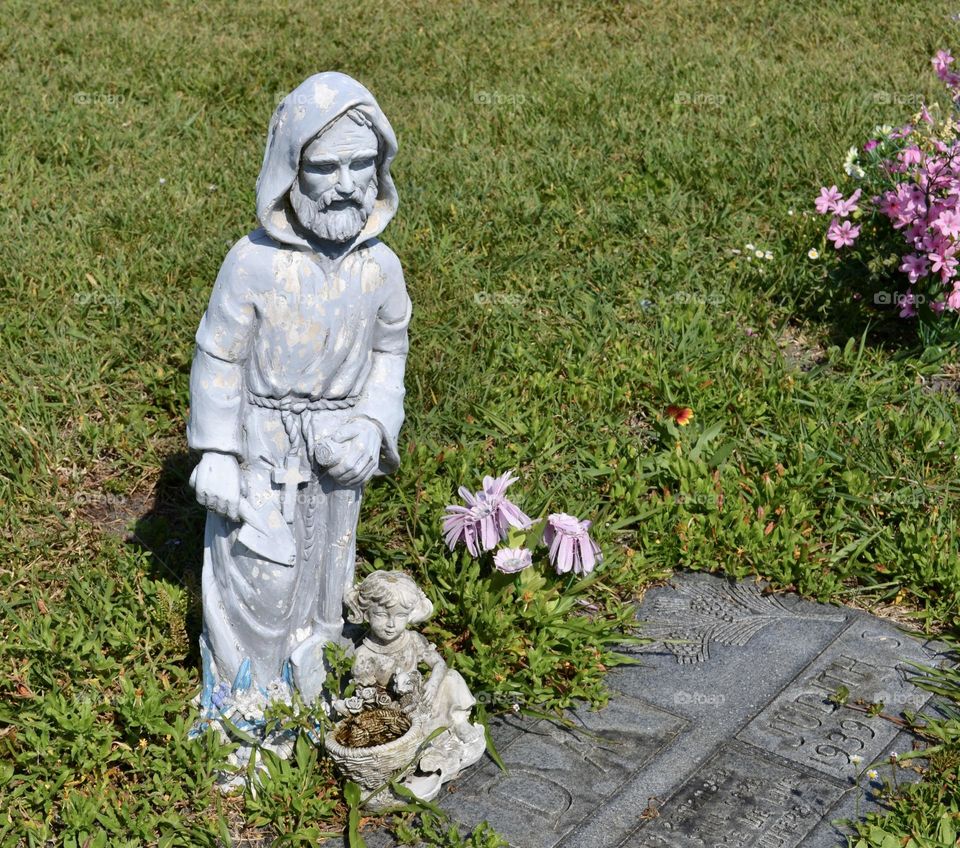 A concrete statue of a man in a hooded robe standing on the ground next to a flat gravestone