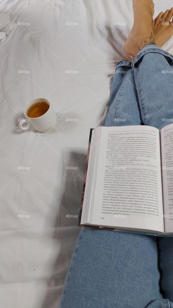 Woman legs with tattoo on bed. Woman reading book and drinking relaxing tea 