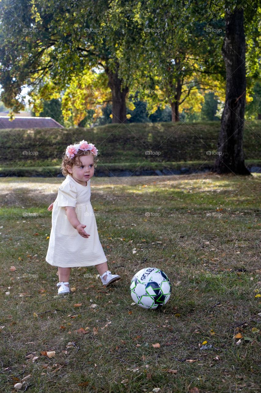 Little fotball girl in action. 
She plays in the park with other Kids.
Dressed in her finest summer dressand flowers in her hair.