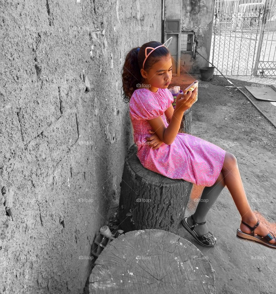 Criança usando vestido rosa, arco de cabeça rosa com orelhas, sandálias pretas, sentada e acessando as redes sociais, através do Smartphone
