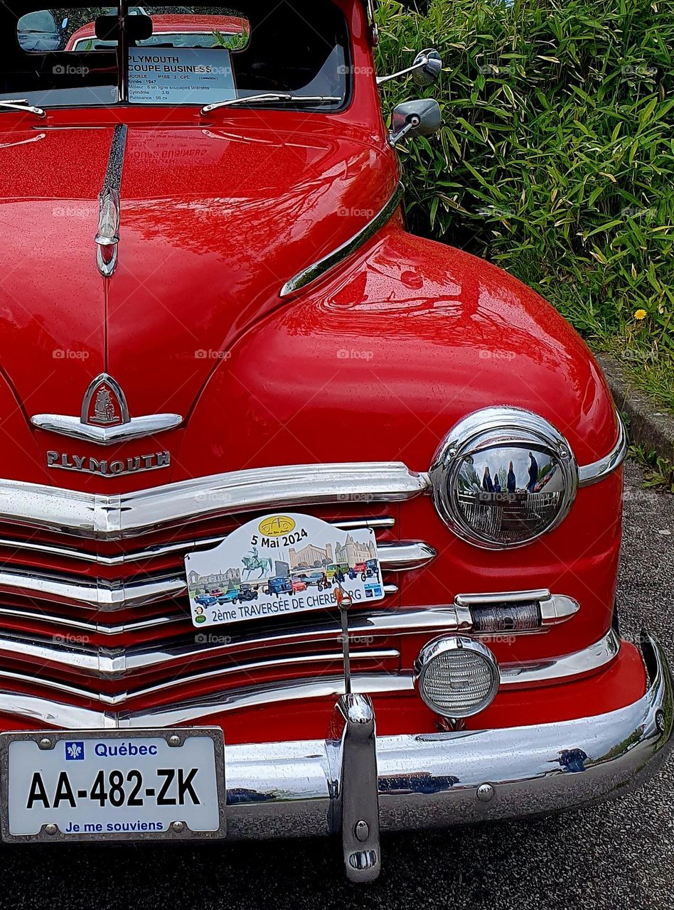 Part of the front of a red Plymouth car showing the grill, one front light and windscreen at Cherbourg car show