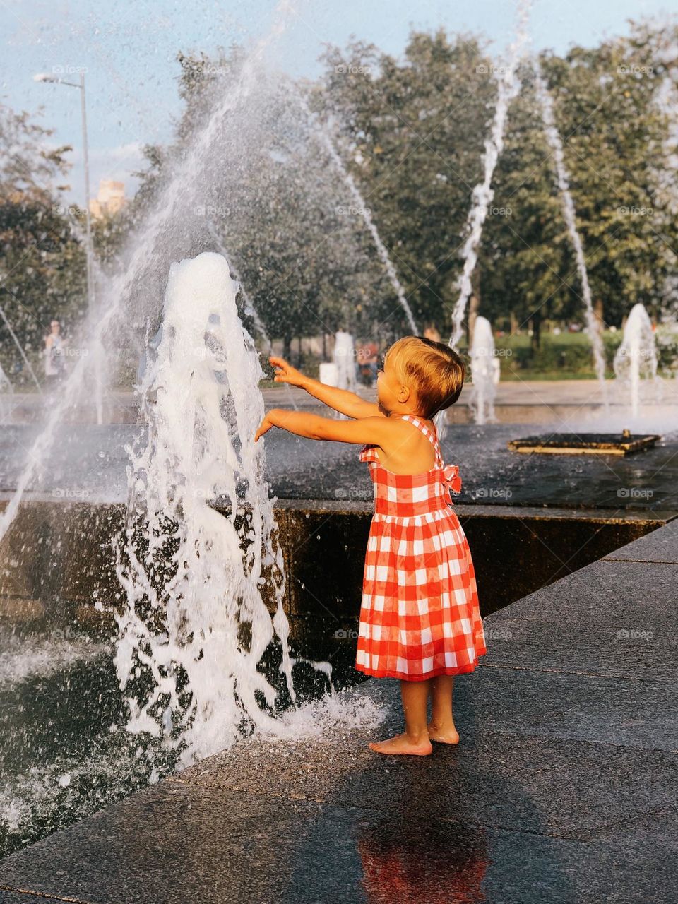 A cute little smiling blonde girl walking near the fountains on a summer sunny day, portrait of child 