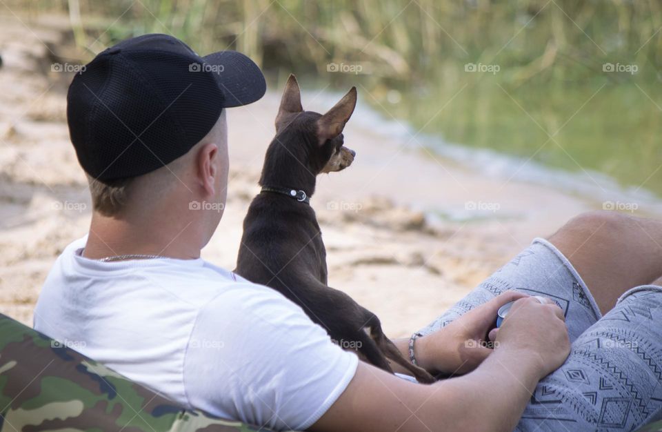 a man sits near the river with a dog,