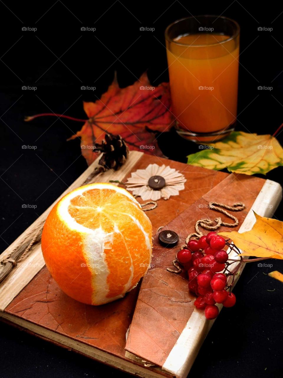 Autumn composition. Book. Half peeled orange and red rowan berries. Next to the book are a red maple leaf, yellow maple leaves. In the background is a glass of orange orange juice. Black background